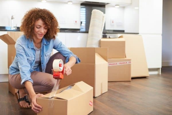 Woman taping moving box closed while surrounded by other moving boxes.