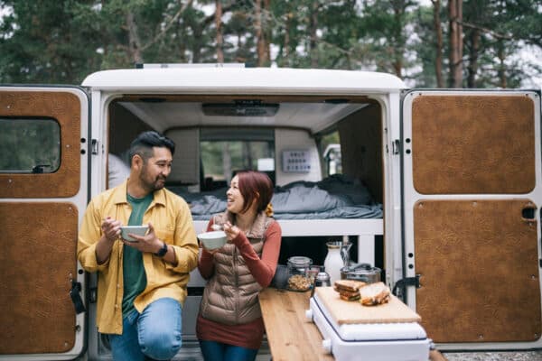 A couple enjoying a meal behind their camping van that is set up with all the doors open.